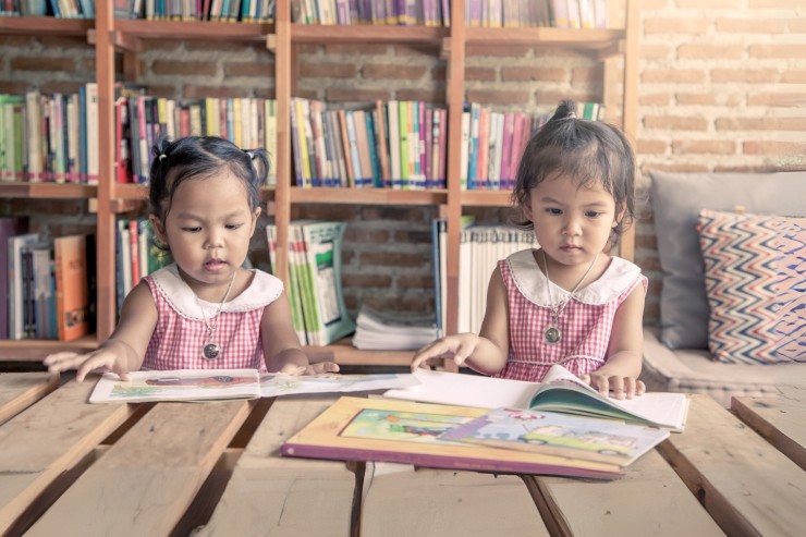 Two young girls in matching dresses sit at a wooden library table reading picture books.