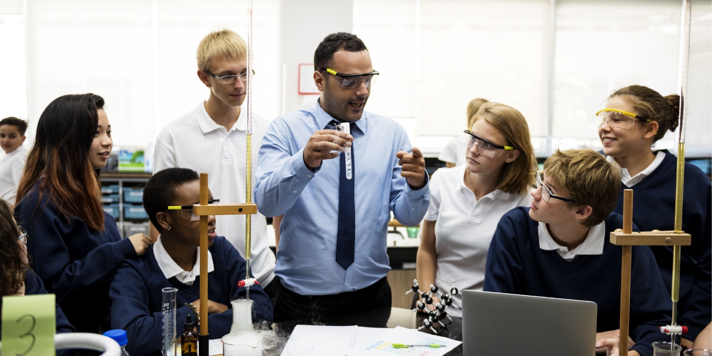 A male teacher in a blue shirt and safety glasses holds up a test tube and looks to be explaining something to a group or students milling around him.