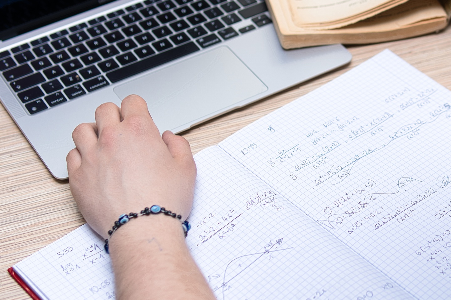 The image shows a teenage boy's hand with beads around the wrist, resting on a page of mathematical equations, with the fingertips on the edge of an open laptop.