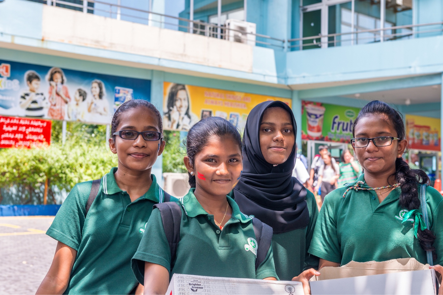 Three female students in green uniform tops stand in the playground of a school in the Maldives, with a woman teacher behind them.