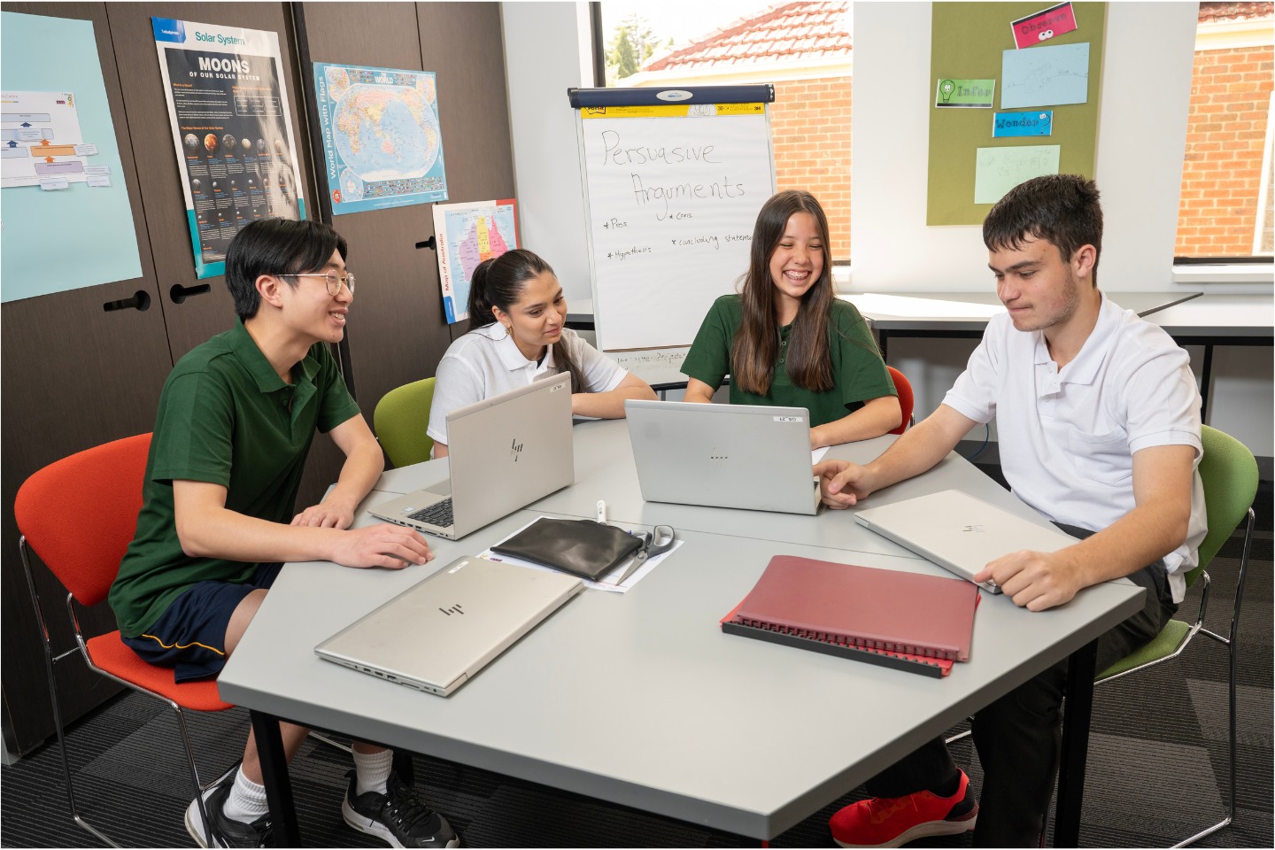 Two teenage boys and 2 teenage girls sit around a table with laptops and workbooks on it. They are happy and looking at one laptop in front of one of the boys.