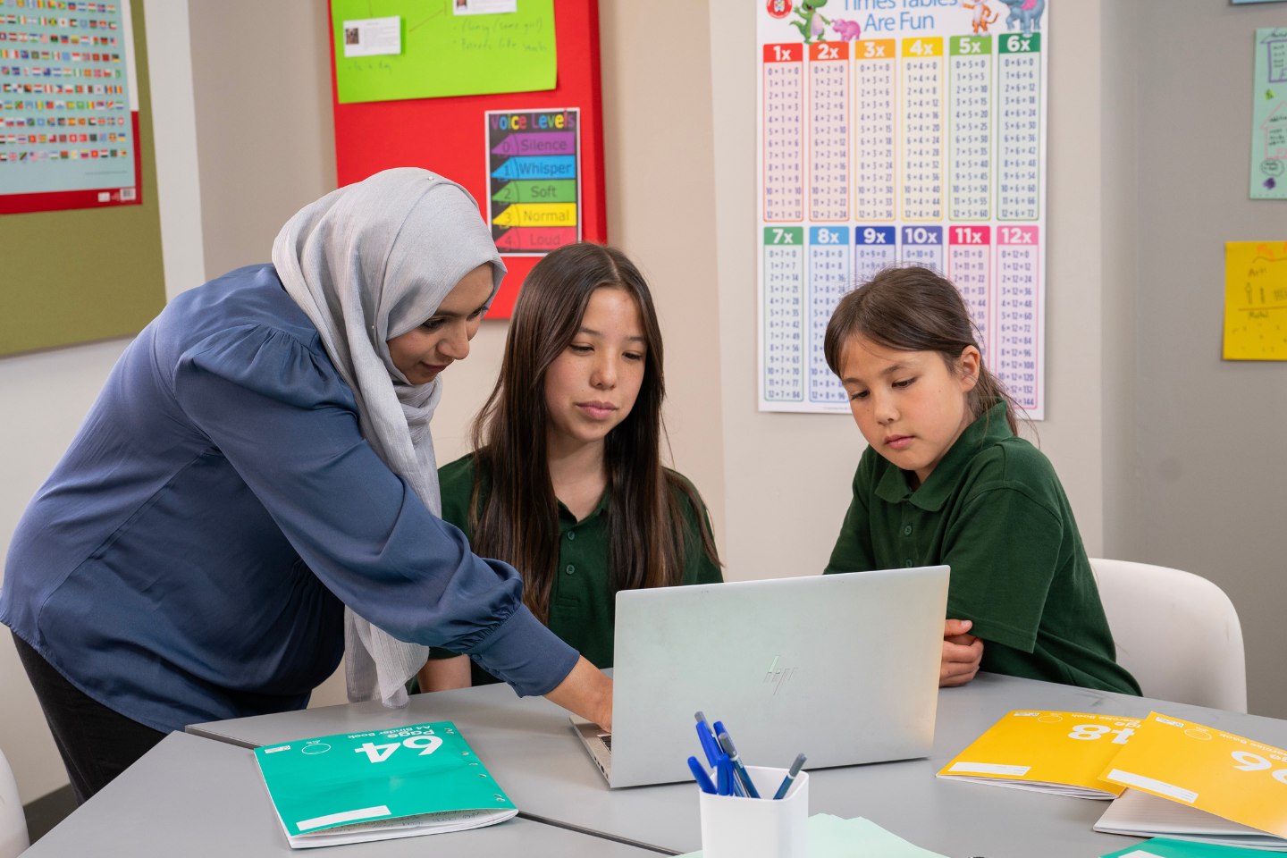 A female teacher leans down to point something out on a laptop between 2 girls.