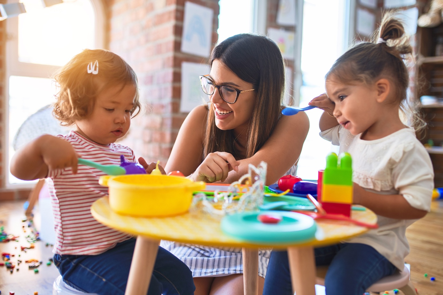 A young female preschool teacher is crouched down with toddlers at a table filled with colourful toys.