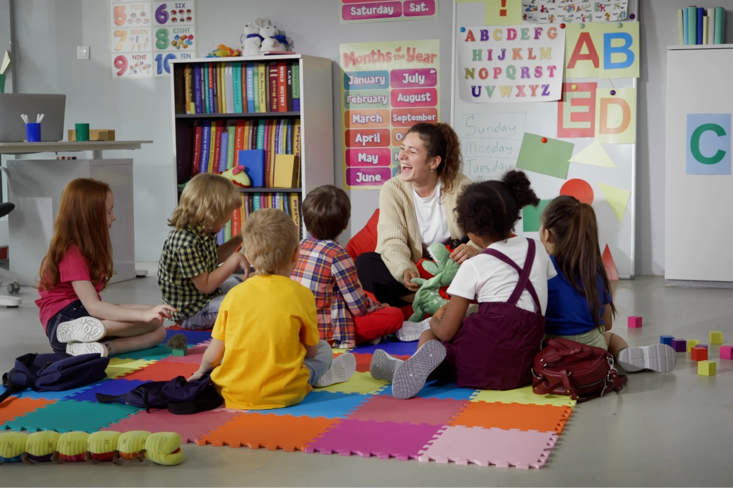 A female teacher sits laughing on the floor of a colourfully decorated kindergarten room surrounded by young children, also sitting.