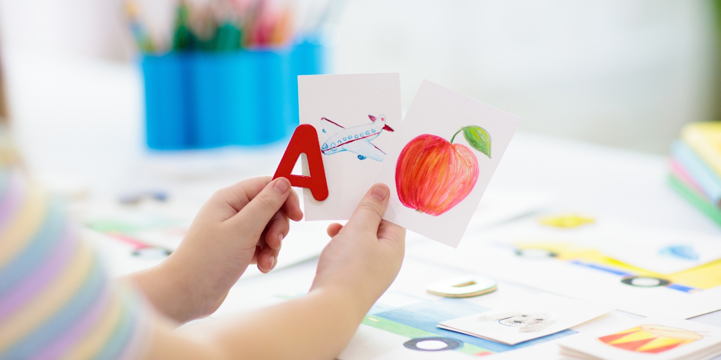 A child's hands are holding the letter A in one and a picture of an apple in the other.  A card with an image of an aeroplane is in between.