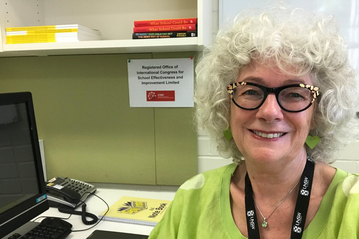 The photo shows a woman - Dr Dudley - from the shoulders up sitting at a desk with a computer on it.  She is smiling and has curly grey hair.