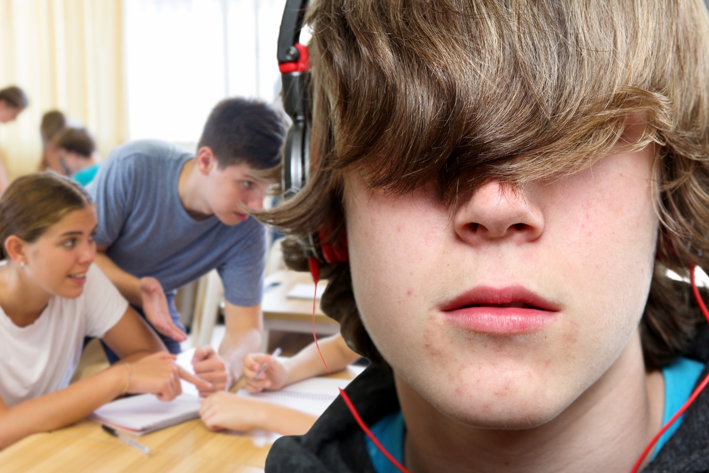 The face of a teenage boy with his hair over his eyes and headphones on is in the foreground. More engaged students collaborating on their work are in the background.