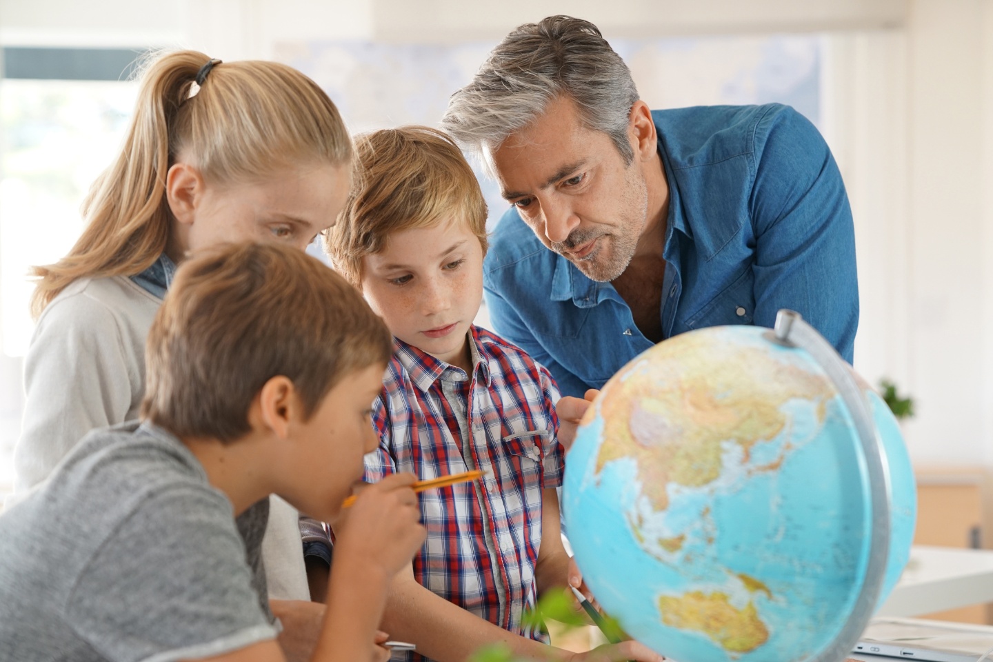 A male teacher points out places on a world globe to a group of young students gathered around it.