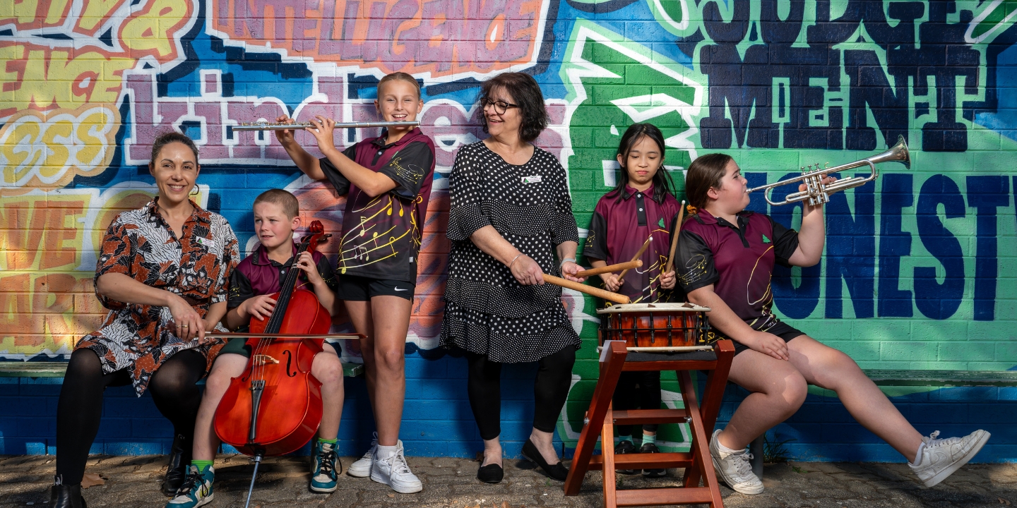 A group of 4 primary school children from The Pines School in Adelaide play instruments - a flute, a cello, a trumpet and a drum - with two teachers, all standing in front of a colourful painted brick wall.