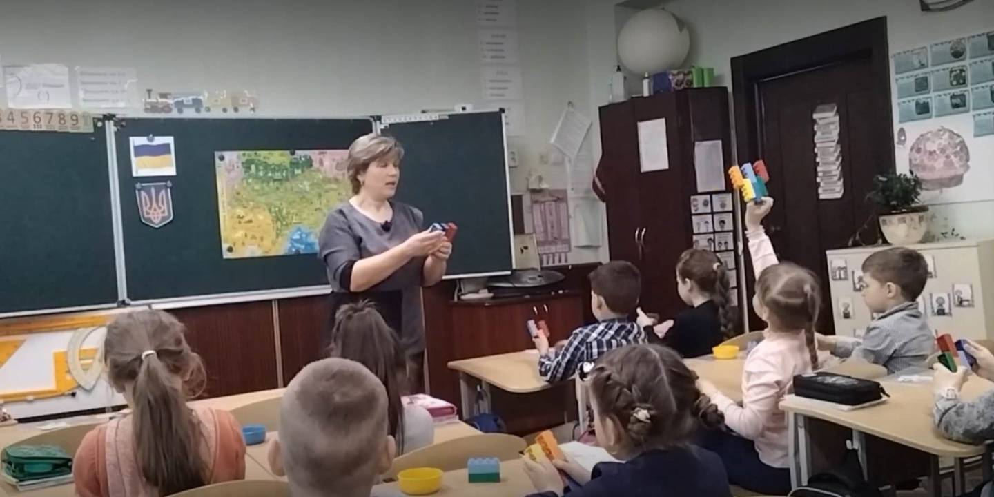A female teacher talks to primary school children sitting at desks with building blocks on them in a Ukrainian classroom. A girl holds her blocks up in the air to show the teacher who has her own block configuration.